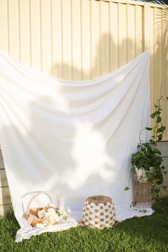 A bedsheet hangs on an iron fench with a basket of flowers, a wicker basket and a pothos plant decorating it in golden hour light